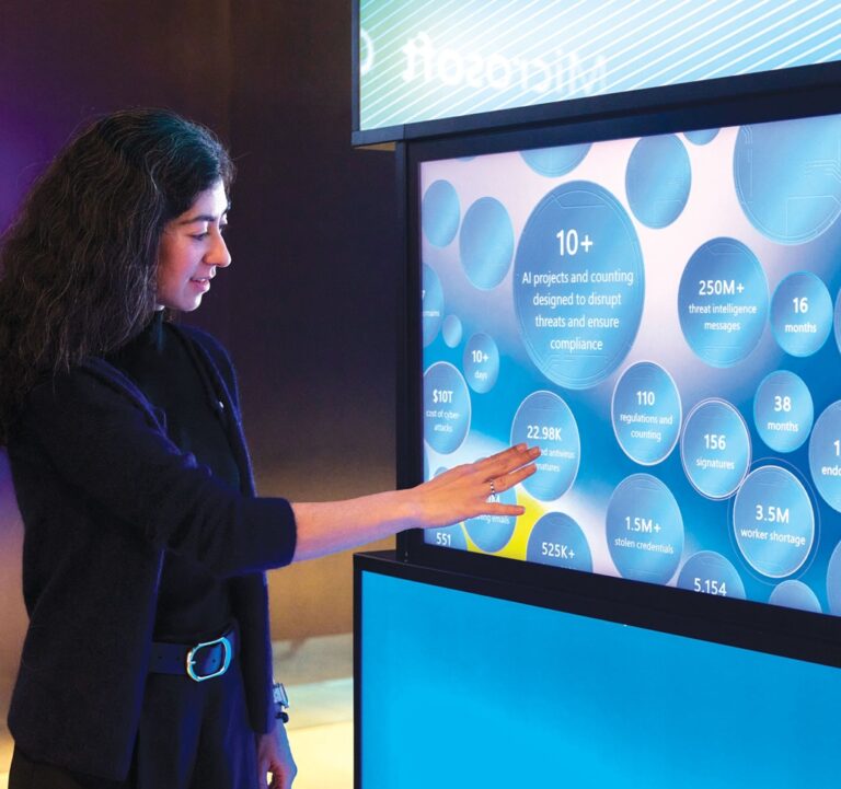 A woman interacts with a large touchscreen display showing data visualizations and statistics related to AI projects and cybersecurity, including circular infographics with numbers and text.